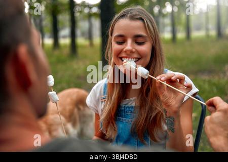 In a joyful moment, a young woman joyfully roasts marshmallows over a glowing campfire in a tranquil forest setting, while sharing laughter and creati Stock Photo