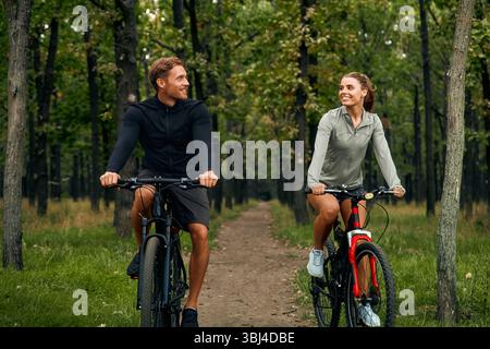 A cheerful couple rides their bicycles together on a beautiful and scenic trail, which is surrounded by lush greenery, while they enjoy a joyful and d Stock Photo