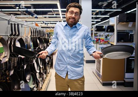 In a bustling kitchenware store, a bewildered man stands holding two frying pans, intensely contemplating his decision as he surveys the wide array of Stock Photo