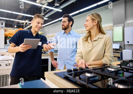 In a modern kitchen showroom, three individuals share engaging interactions as they showcase various kitchen appliances while enjoying a friendly conv Stock Photo