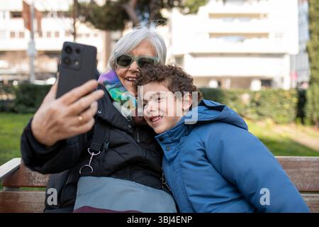 Grandmother and grandchild are sitting on a park bench taking a selfie with a smartphone and smiling Stock Photo