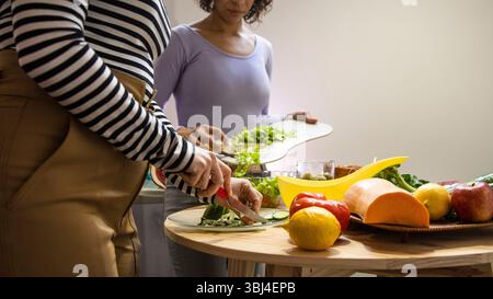 Two people chop and arrange fresh vegetables on a wooden table in a warm kitchen, enjoying their cooking time together. Stock Photo