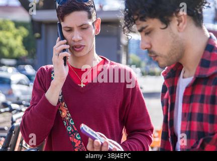 Two young men are engaged in conversation outdoors, with one on the phone while holding a smartphone. Stock Photo