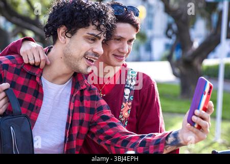 Two young men are smiling and posing for selfies in a park on a sunny day. They seem to be having a great time together. Stock Photo