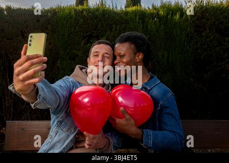 Happy interracial gay couple taking a selfie with heart balloons in a park, celebrating valentine's day Stock Photo