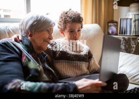 Grandmother and grandchild enjoying time together, using a laptop on the sofa in a cozy home setting, sharing a special moment of connection Stock Photo