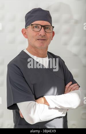 Male dentist wearing scrubs and glasses stands with crossed arms, exuding confidence and professionalism in a modern dental clinic Stock Photo