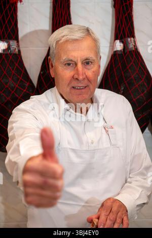 Senior butcher giving a thumbs up while skillfully slicing serrano ham in his bustling shop, radiating pride and expertise Stock Photo