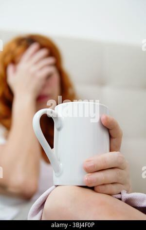 Woman with red hair holding a mug and suffering from strong headache while resting on a sofa Stock Photo