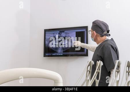 Dentist wearing surgical mask and uniform pointing at teeth x ray displayed on a digital screen in a dental clinic Stock Photo