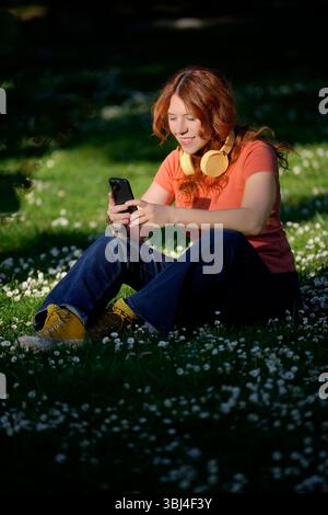 Smiling redhead woman using mobile phone in a field of daisies with headphones around her neck Stock Photo