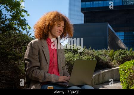 Young man with curly hair working on a laptop outdoors, surrounded by greenery and modern architecture. Concept of sustainable remote work, urban well Stock Photo