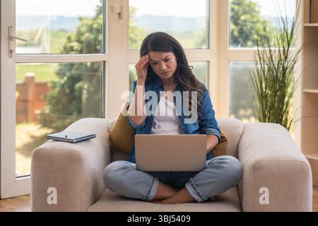 Young businesswoman working remotely from home, sitting on a cozy armchair with a laptop. Modern workspace, digital job, comfortable environment, and Stock Photo