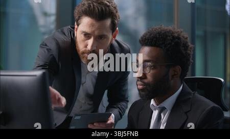 Two multinational businessmen African American Caucasian men diverse males looking pointing at computer monitor pc guys discuss project team working Stock Photo