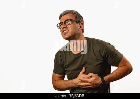 A man is experiencing discomfort while standing against a light background. He holds his chest with one hand, showing a pained expression. Stock Photo
