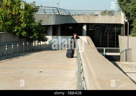 Young woman pulling wheeled luggage while walking up a wide concrete path. Urban image representing travel, movement and independence. Stock Photo