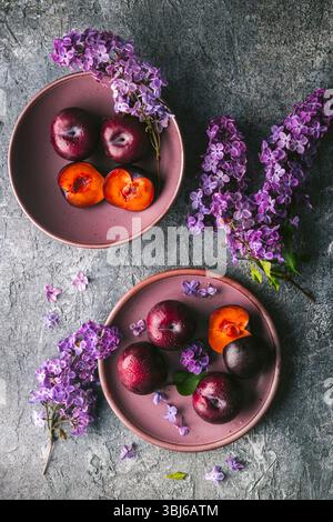 Two mauve-colored bowls on a custom grey, textured surface with an arrangement of several ripe plums, two cut in half to reveal the flesh. Lilac flowe Stock Photo