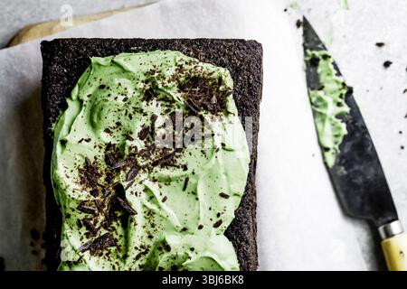 A chocolate cake topped with green frosting and chocolate shavings on a white surface with a knife nearby. Stock Photo