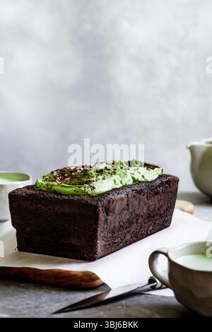 A chocolate loaf cake topped with green frosting and chocolate shavings, placed on a wooden board with a knife and a cup nearby. Stock Photo