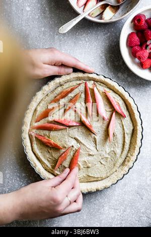 A person decorates a pie with rhubarb slices on a light gray countertop, with bowls of rhubarb and raspberries nearby. Stock Photo