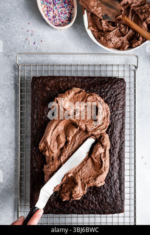 A person spreads chocolate frosting on a rectangular cake with a spatula, next to bowls of frosting and sprinkles. Stock Photo