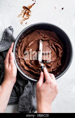 A person spreads chocolate frosting in a cake pan with a spatula on a light surface with a gray cloth nearby. Stock Photo