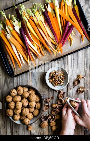 An assortment of colorful carrots on a baking tray alongside a bowl of walnuts and a person cracking open walnuts on a wooden table. Stock Photo