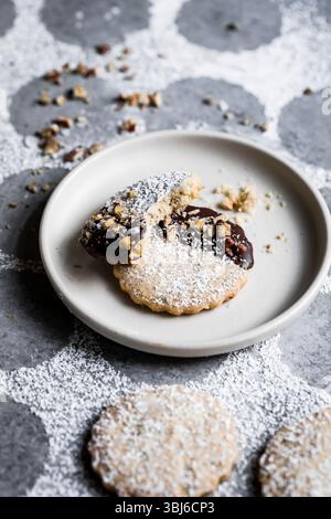 A plate with two cookies, one partially dipped in chocolate and sprinkled with chopped nuts, dusted with powdered sugar. Stock Photo