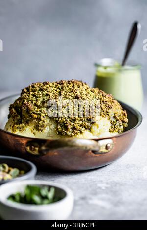 A roasted cauliflower head coated with a green crust, served in a copper pan with a creamy dip in the background. Stock Photo