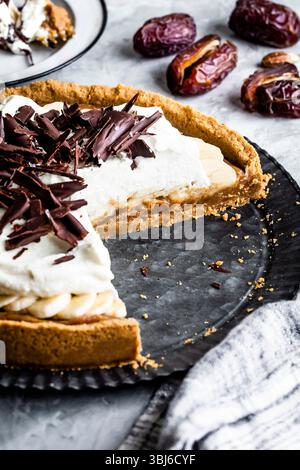 A sliced banoffee pie topped with whipped cream and chocolate shavings, displayed on a metal tray with dates in the background. Stock Photo