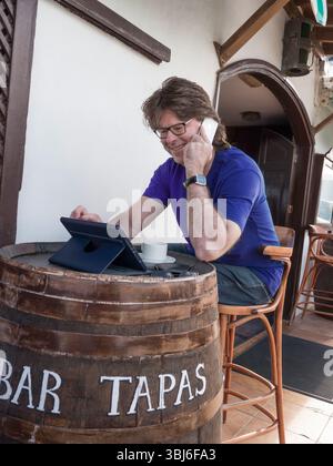 Mature businessman using his iPhone smartphone & iPad Air tablet computer abroad alfresco Spanish Tapas restaurant bar, international communications. Stock Photo