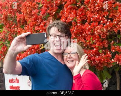 'SELFIE' Attractive couple having fun on holiday taking a 'selfie' photo outside holiday attraction on their iPhone smartphone camera application Stock Photo
