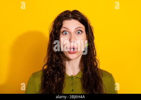 Surprised young woman with curly brown hair on a vibrant yellow background wearing fashionable green apparel Stock Photo