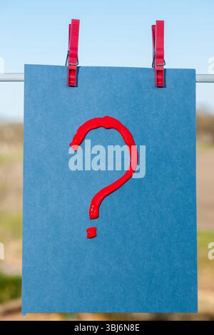 Red question mark painted with acrylic paint on blue cardboard with two clothespins attached to a clothesline against a rural background. Stock Photo