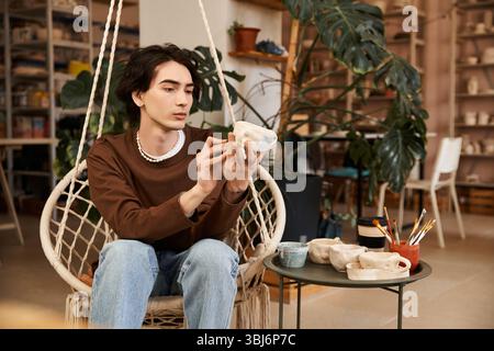 A stylish young man carefully examines his pottery creation in a vibrant studio brimming with art. Stock Photo