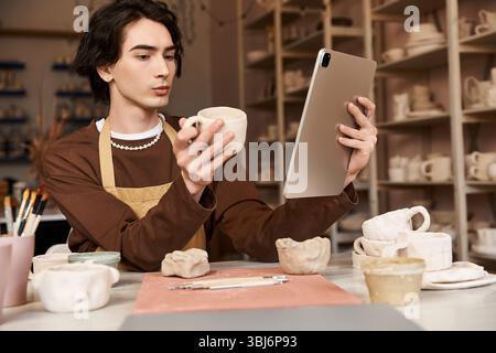 A man in a pottery studio examines a clay mug while using a tablet for creative inspiration. Stock Photo