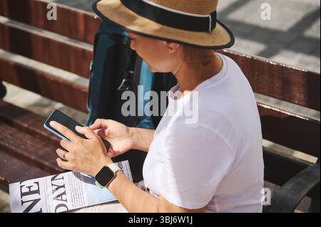 Female traveler wearing casual clothes and a hat sitting on a bench, using her smartphone, and enjoying the sunny outdoors during a summer day. Stock Photo