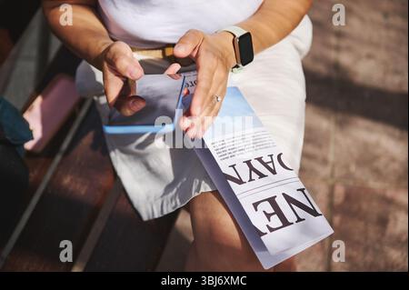 A woman interacts with her smartphone while holding a folded newspaper on a sunny day. She is outdoors, showcasing casual attire and focus on her hand Stock Photo