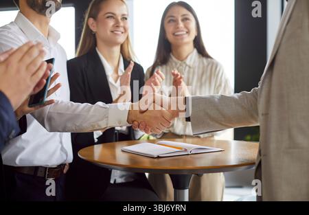 Two business professionals shaking hands while their colleagues celebrating successful partnership. Stock Photo