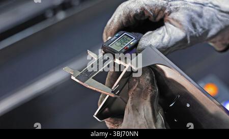Worker measuring metal part with digital caliper. A gloved hand is using a digital caliper to precisely measure the width of a metallic component. Stock Photo