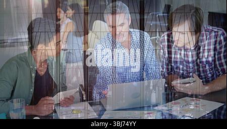 Three men using laptop tablet checking smartphone amid printed charts, glass reflection in office Stock Photo