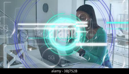 Holding mug, mid adult woman typing on silver laptop at open-plan office, with 5G interface Stock Photo