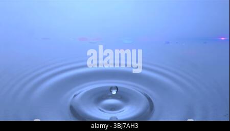 Capturing clear water droplet forming ripples on blue surface in macro setup, with lens flare Stock Photo