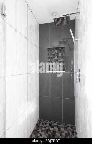 A modern shower with large format white and dark grey tiles, mosaic square tile flooring and shelf, and a chrome showerhead. Stock Photo