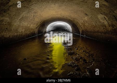 Tunnel of the old semicircular sewer drainage tunnel with gaps in the middle, from which comes a yellow light. Stock Photo