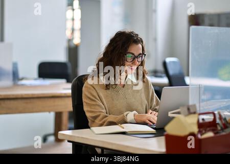 Young happy professional business woman sitting at desk working on laptop. Stock Photo