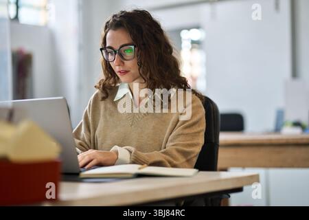 Young happy woman employee or student sitting at desk using laptop. Stock Photo