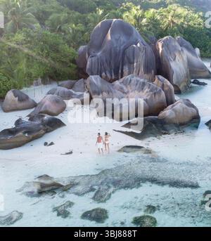 Aerial view of young couple walking on dream beach on Seychelles Island. Woman and man are holding hands in front of unique rock formations on island. Stock Photo