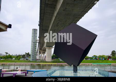 Incheon, South Korea - May 19th 2025: A tilted black cube sculpture rests on a glass platform beneath the Ara Waterway’s Gyeyang Bridge, flanked by el Stock Photo