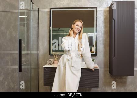 Pretty woman wearing white bathrobe, posing near sink and mirror in bathroom Stock Photo